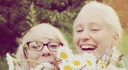 Zwei junge Frauen lachen in die Kamera, halb verdeckt von einem Margeritenstrauß.